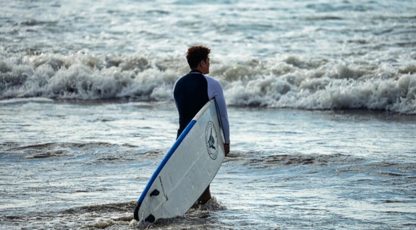 A surfer holding a board stands in the ocean waves at a Bali beach, showcasing a perfect sunset scene.