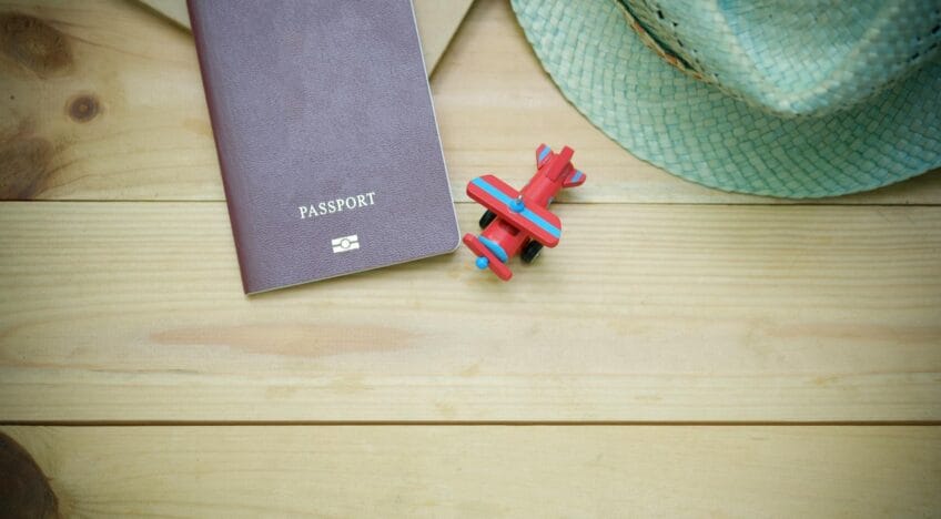 Flat lay of a passport, straw hat, and toy airplane on a wooden surface.